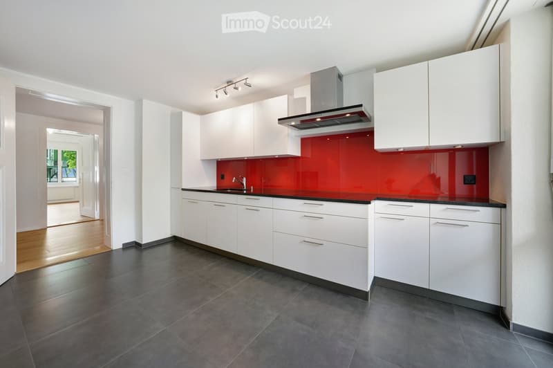 Modern kitchen with white cabinets, dark countertops, and a red backsplash. Features a stainless steel range hood and a dark tiled floor.