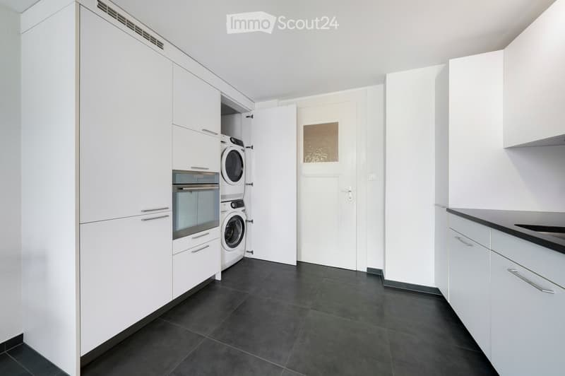 Modern kitchen with white cabinets, black countertop, and built-in appliances including a washing machine and oven. The space features dark tiled flooring and a door leading to another area.