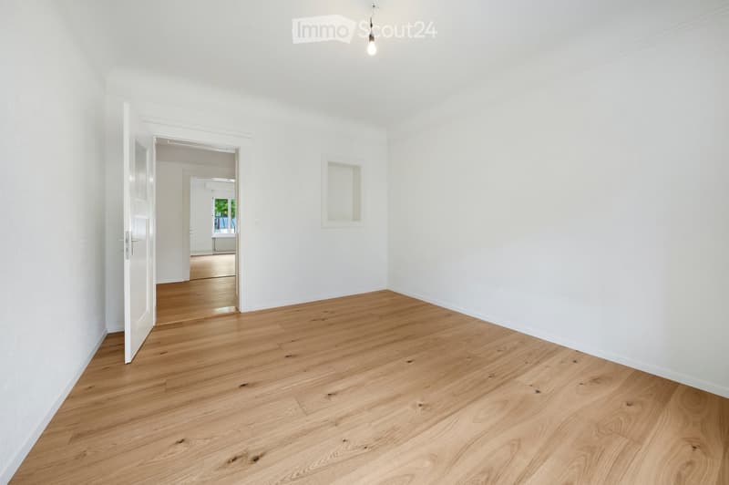 Empty room with light wooden flooring and white walls, featuring an open doorway leading to another room.