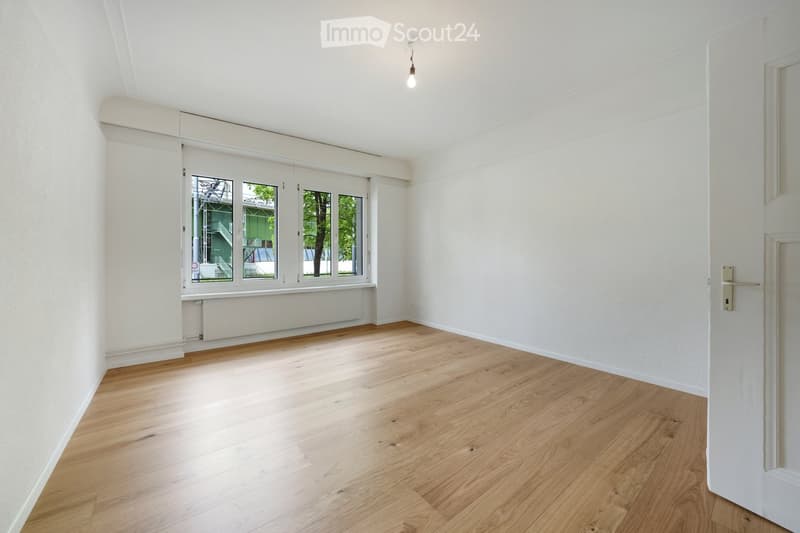 Empty room with light wooden flooring, white walls, and large windows with blinds. The room has a radiator under the windows and a single light fixture on the ceiling.