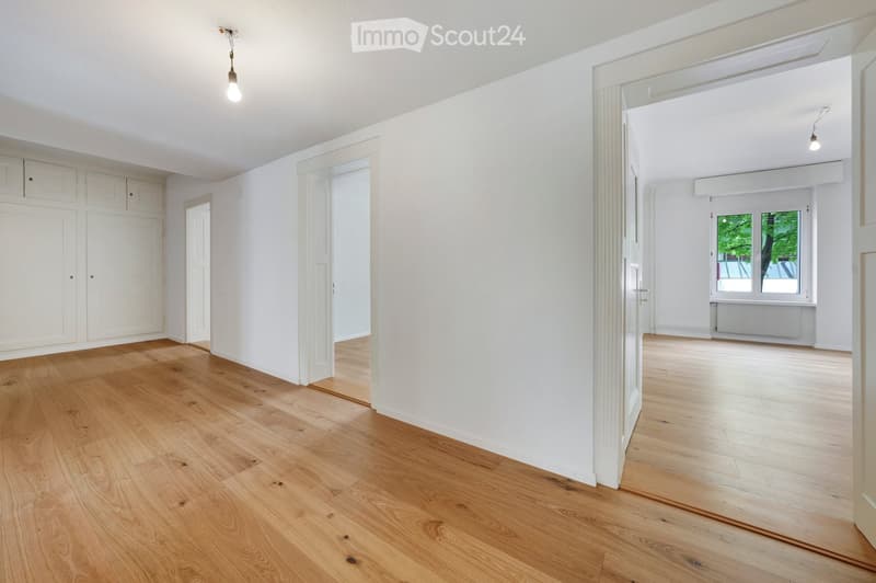 Empty room with light wooden flooring and white walls, featuring built-in cabinets and a doorway leading to another room with a window and radiator.