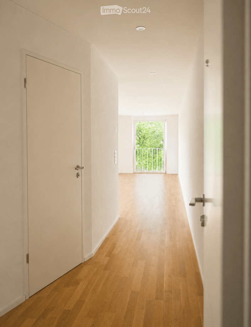 A modern, minimalist hallway with light wooden flooring, white walls, and recessed ceiling lighting. Features a closed door on the left and an open doorway leading to a balcony or outdoor area with greenery visible beyond.