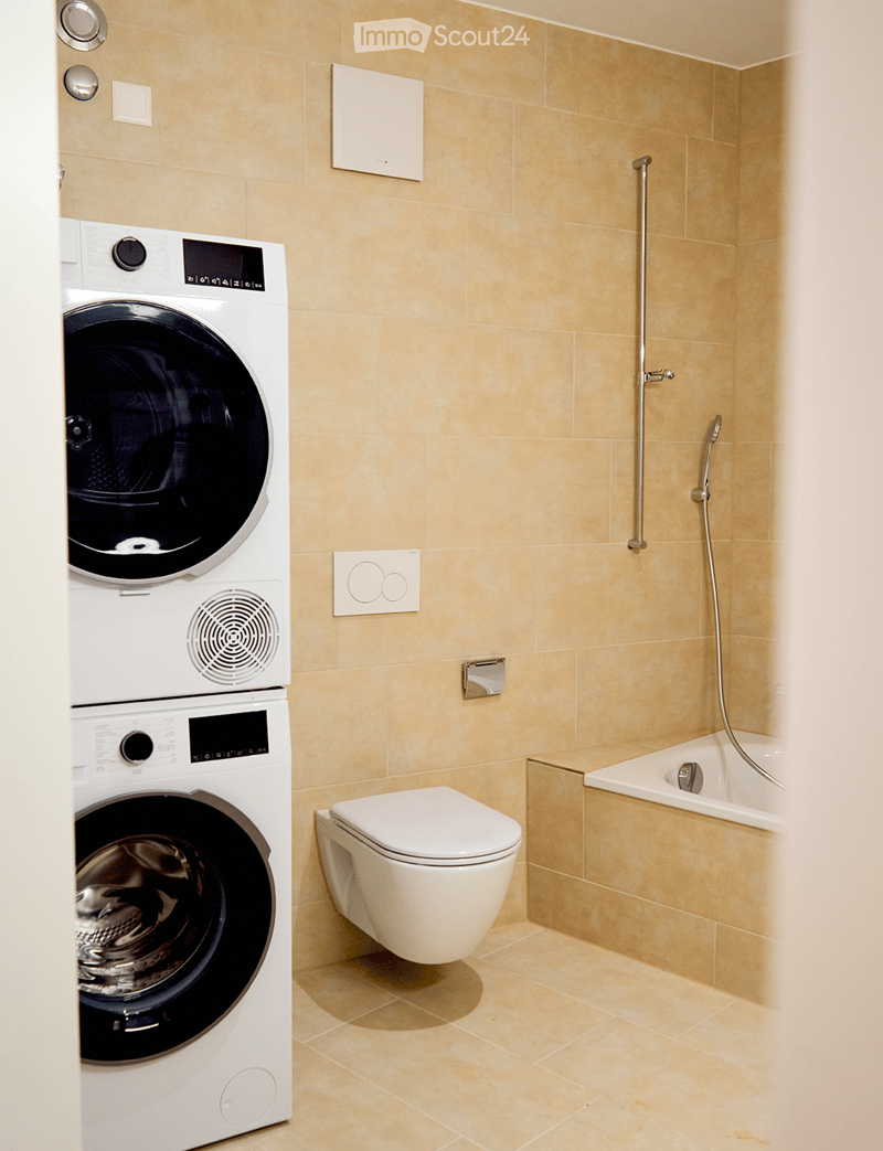 Modern bathroom with beige tiles, a white toilet, a bathtub with a shower hose, and a stacked washing machine/dryer unit.