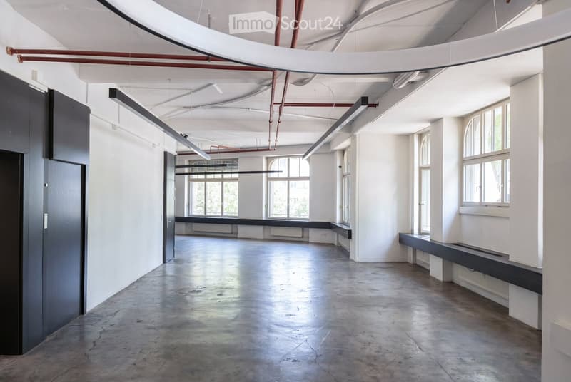 An empty, industrial-style room with white walls, concrete floor, large windows, exposed pipes, and built-in benches along the walls.