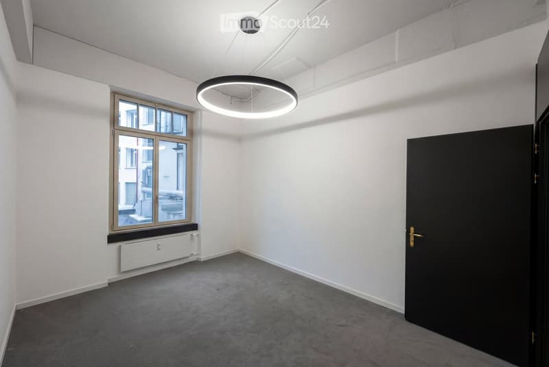 Empty room with white walls, grey carpeted floor, large window with wooden frame, and a modern circular pendant light fixture.