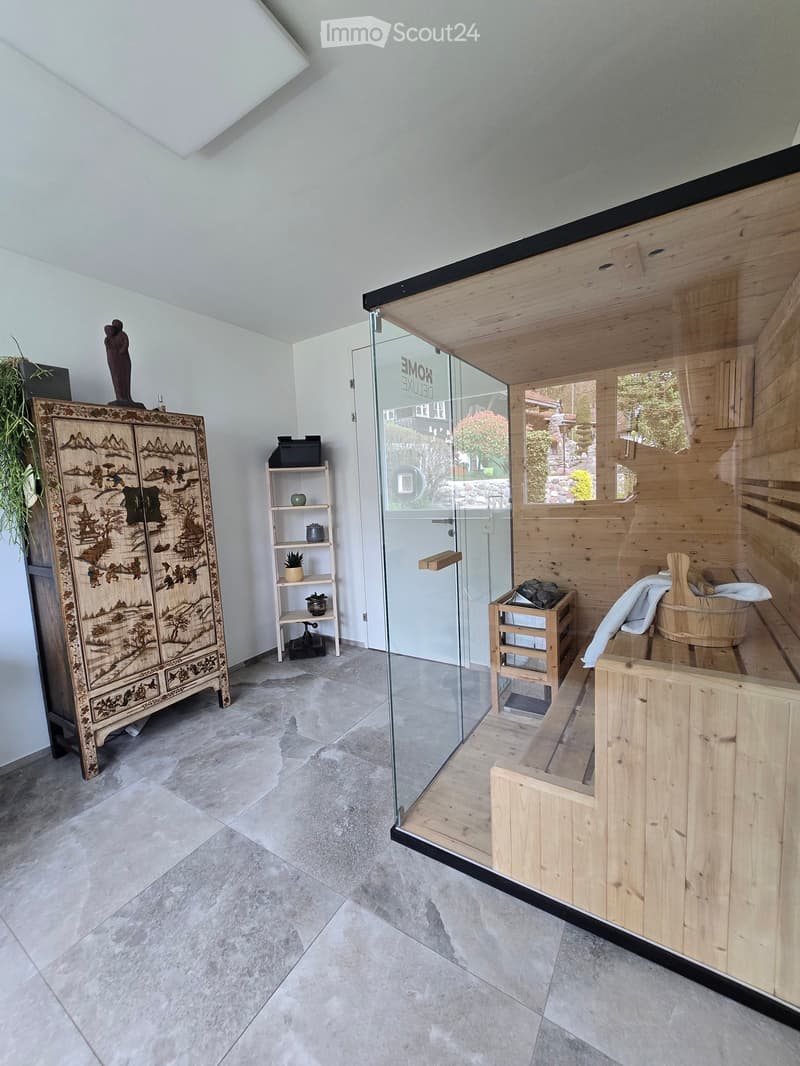 A modern, well-lit room with a wooden sauna area enclosed by glass, featuring a wooden bench, a wooden tub, a cabinet with intricate carvings, a small shelf with decorative items, and a hanging plant.