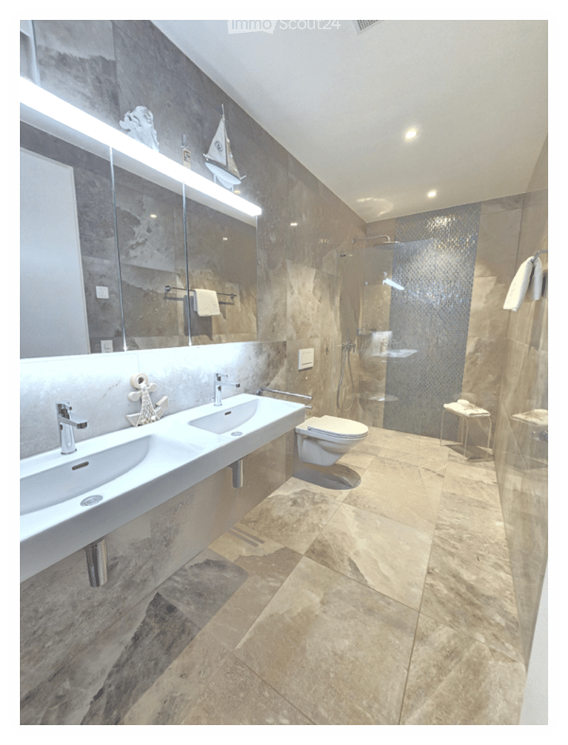 Modern bathroom with double sinks, a large mirror, a toilet, a glass-enclosed shower, and a small stool with towels.