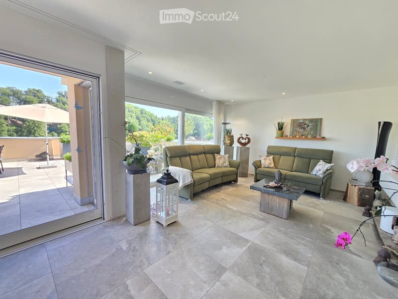 Bright living room with large windows and a view of greenery, featuring two green sofas, a coffee table, decorative vases, and artwork.