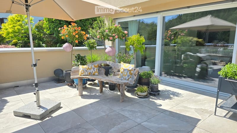 A sunny outdoor patio with a wooden bench, colorful cushions, potted plants, and a large umbrella. The space features tiled flooring and a view of greenery through large glass doors.