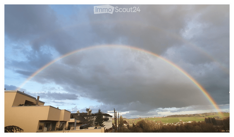A view of a landscape with a double rainbow in the sky, featuring a house and trees in the foreground.