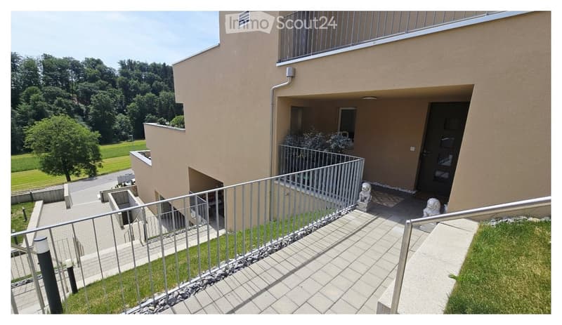 Balcony with metal railing, tiled floor, small garden area, and view of a building entrance and surrounding greenery.