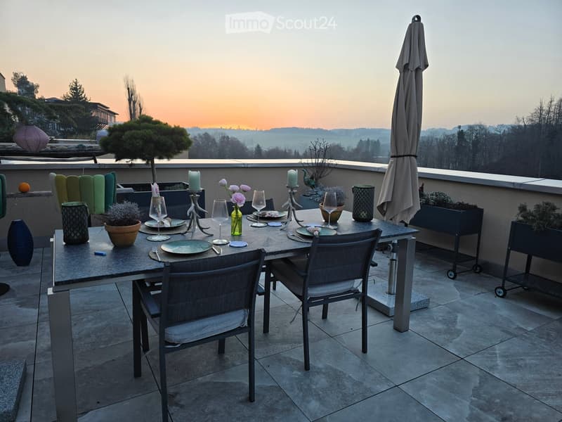 A well-set dining table on a balcony or terrace with a scenic view of a valley at sunset. The table is set with plates, wine glasses, candles, and decorative vases. There are chairs around the table and an umbrella nearby.