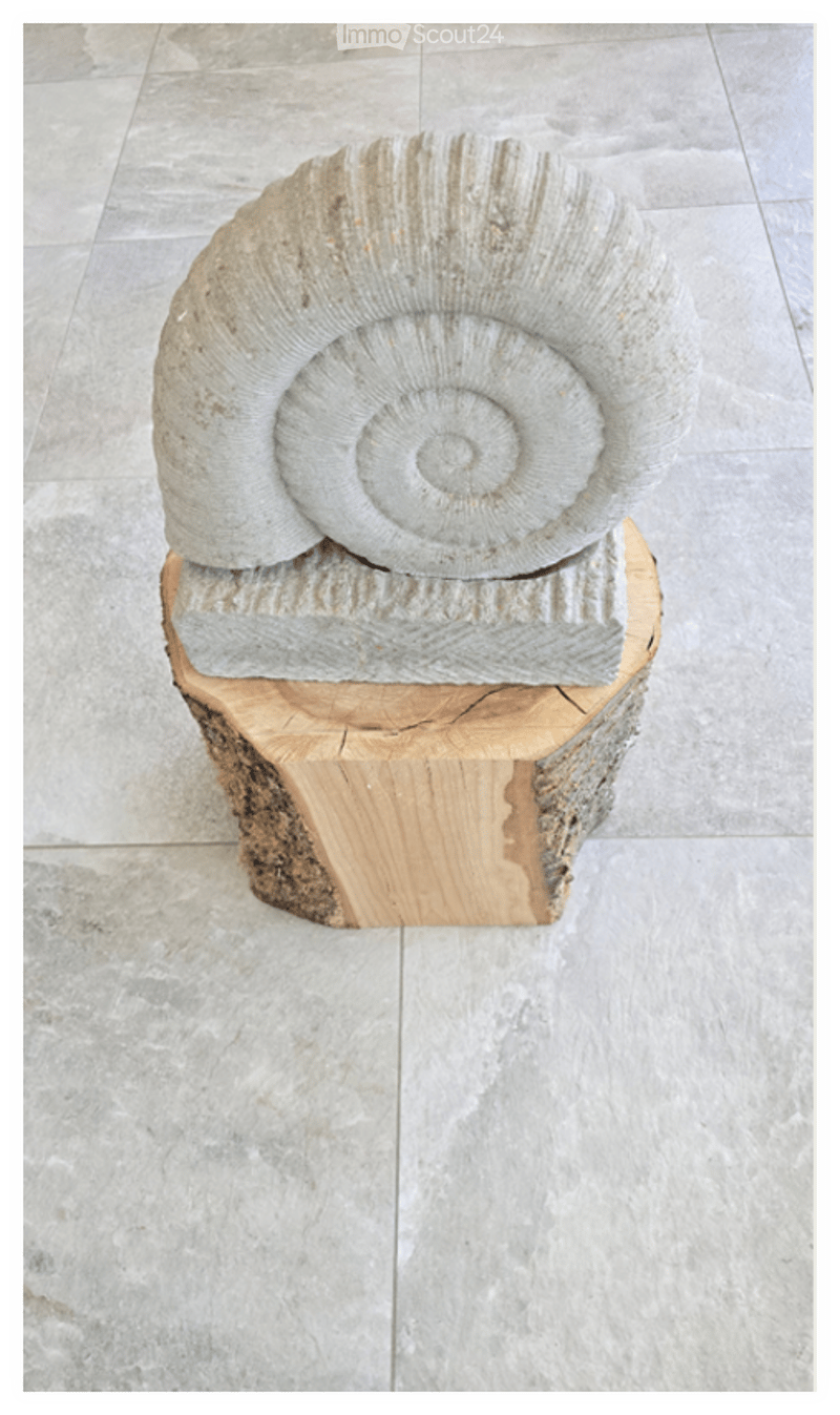 A large, spiral-shaped stone sculpture is displayed on a wooden base in a room with a tiled floor.