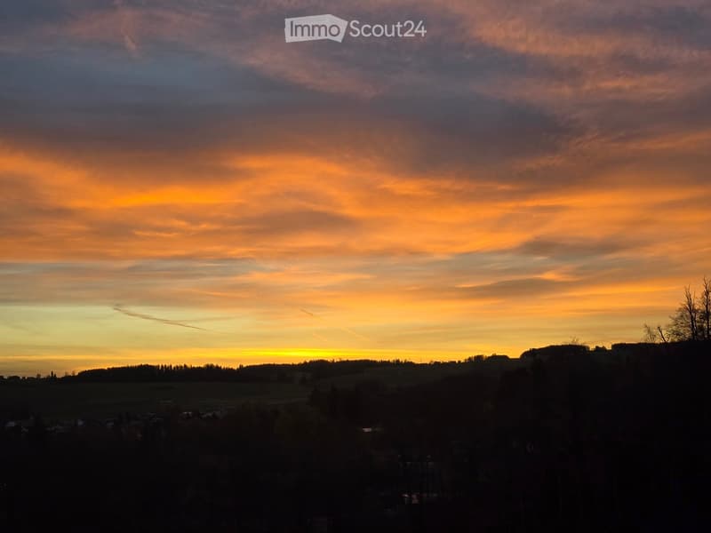 A panoramic view of a rural landscape at sunset, with rolling hills, scattered trees, and a vibrant orange sky.