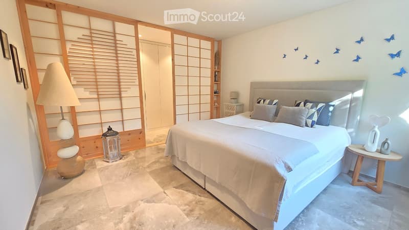 Modern bedroom with a large bed, nightstand, table lamp, decorative vase, and a decorative lantern. The room features a sliding wooden door and a tiled floor.