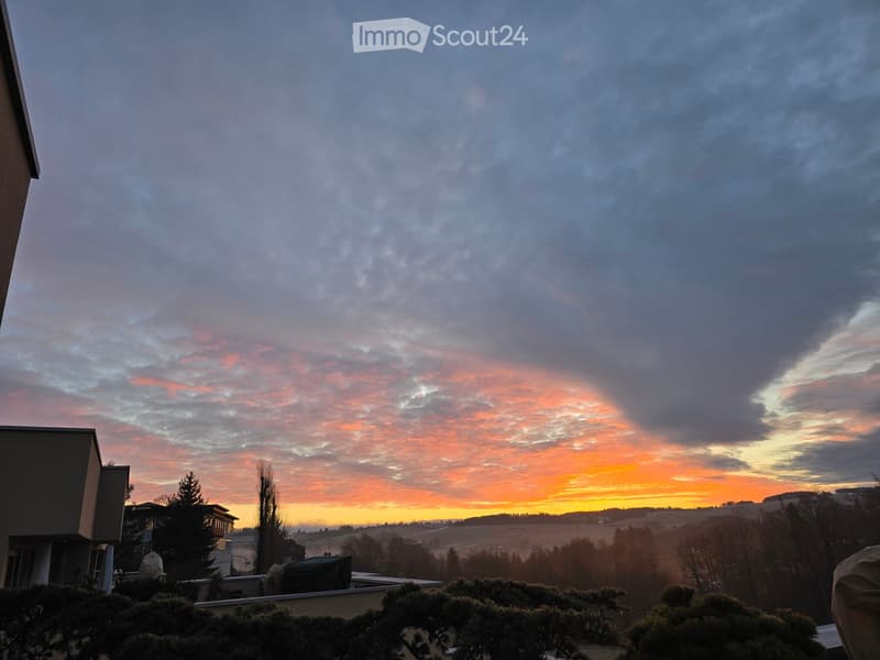 Balcony view of a sunset over a rural landscape with fields, trees, and distant hills.