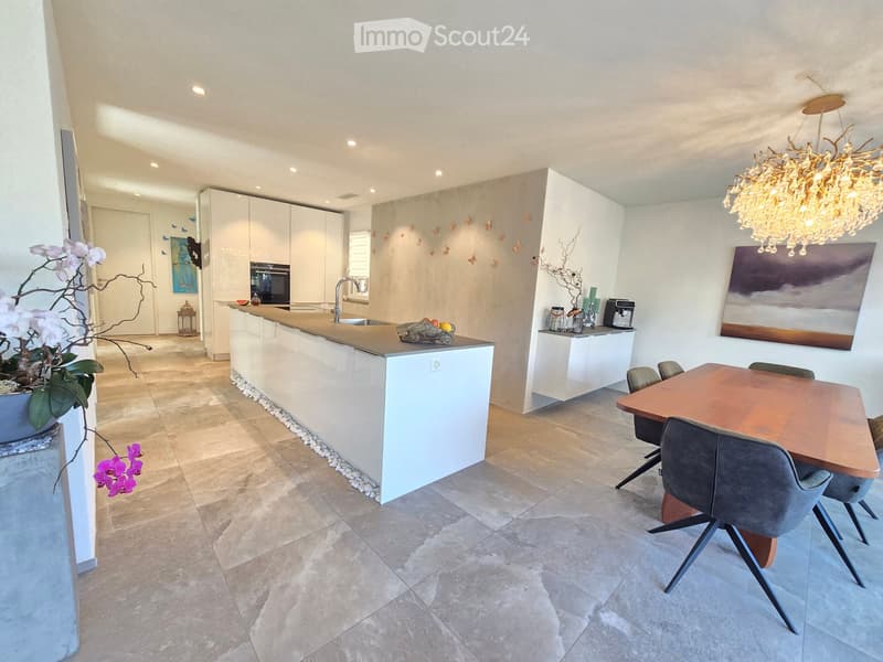 Modern kitchen with white cabinets, a central island, a dining table with chairs, and a chandelier. Features a sink, faucet, coffee maker, and decorative artwork.