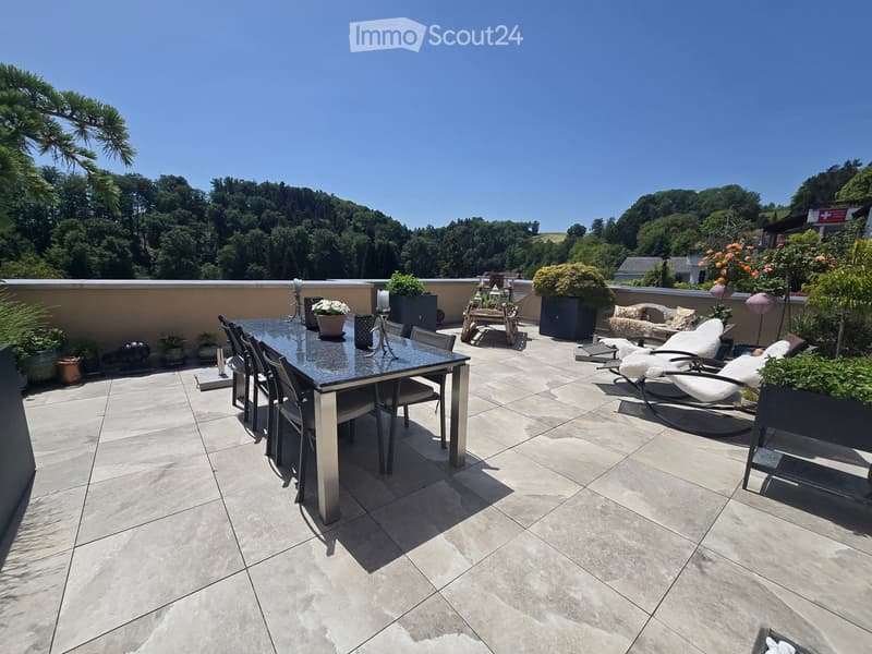 A spacious balcony with a tiled floor, a dining table with chairs, lounge chairs, and potted plants, offering a scenic view of greenery.