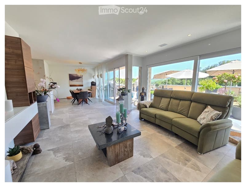 Modern living room with large windows offering a view, featuring a green sofa, coffee table, dining area, chandelier, and decorative items.
