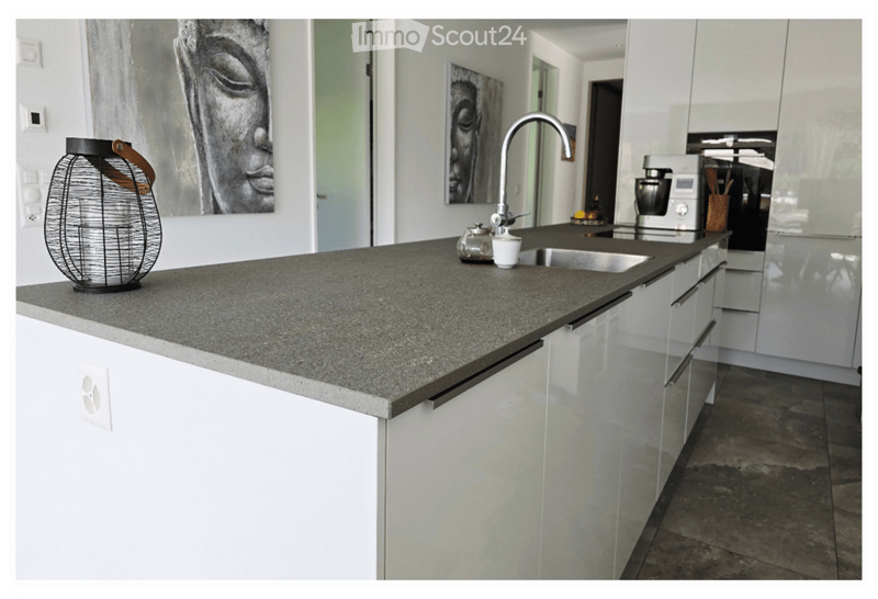 Modern kitchen with white cabinets, a grey granite island, a stainless steel sink, and various kitchen appliances. Decorative items and artworks are placed on the island and walls.