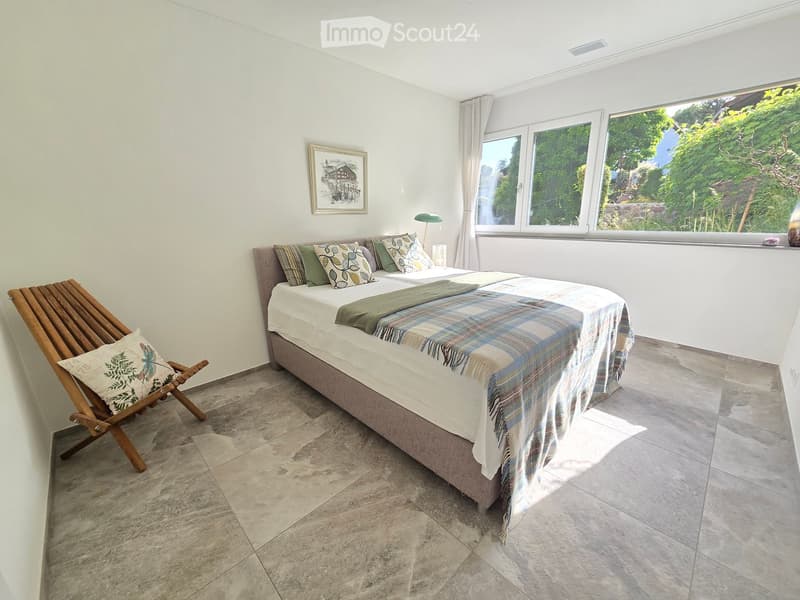 Bright bedroom with a large bed, colorful pillows, plaid blanket, wooden chair, and a view of greenery through large windows.