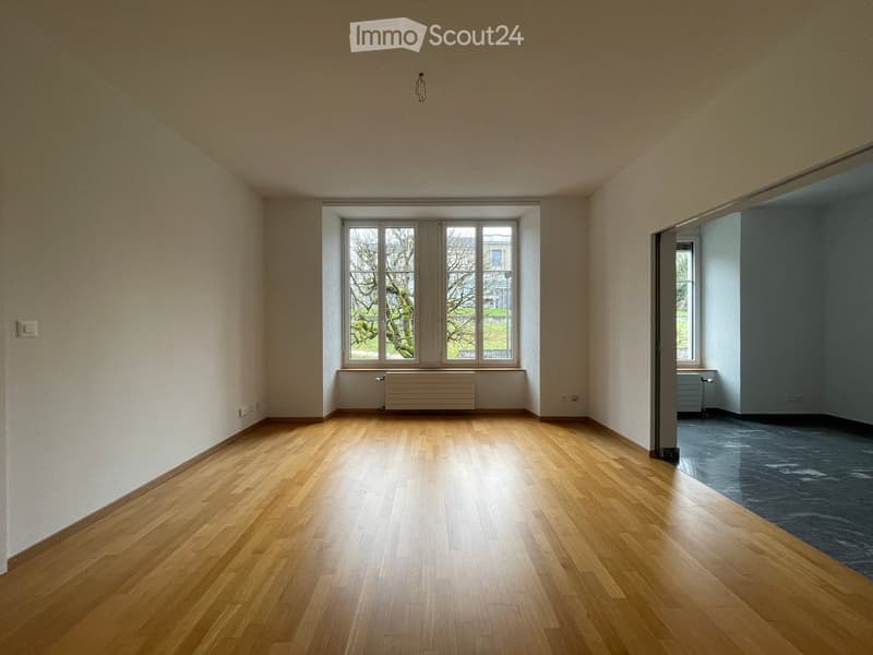 Empty room with wooden flooring, large window with white frame, radiator below window, and adjacent room visible through doorway.