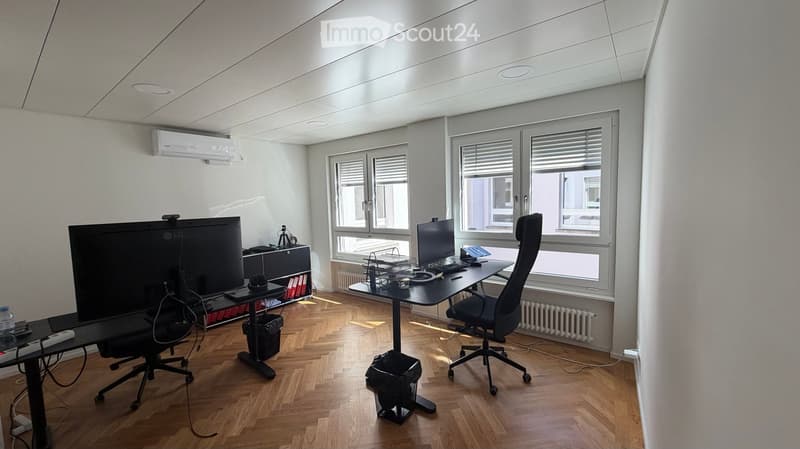 Office space with wooden flooring, large windows, two desks with chairs, multiple screens, laptops, and office supplies.