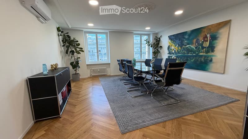 Modern furnished room with wooden flooring, large windows, a grey rug, a black cabinet, a large painting, and a set of black chairs around a glass table.