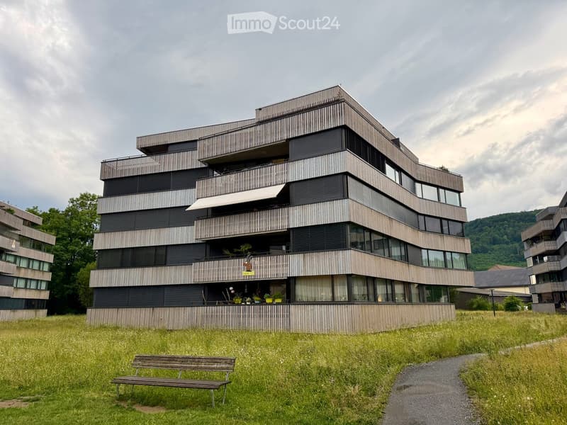 Modern multi-story residential building with balconies, large windows, and a sleek design, surrounded by a grassy area with a bench.
