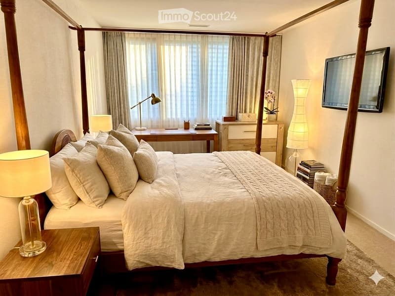 Elegant bedroom with a four-poster bed, nightstand, desk, and large window with curtains. Features include a TV mounted on the wall, a floor lamp, and decorative items.