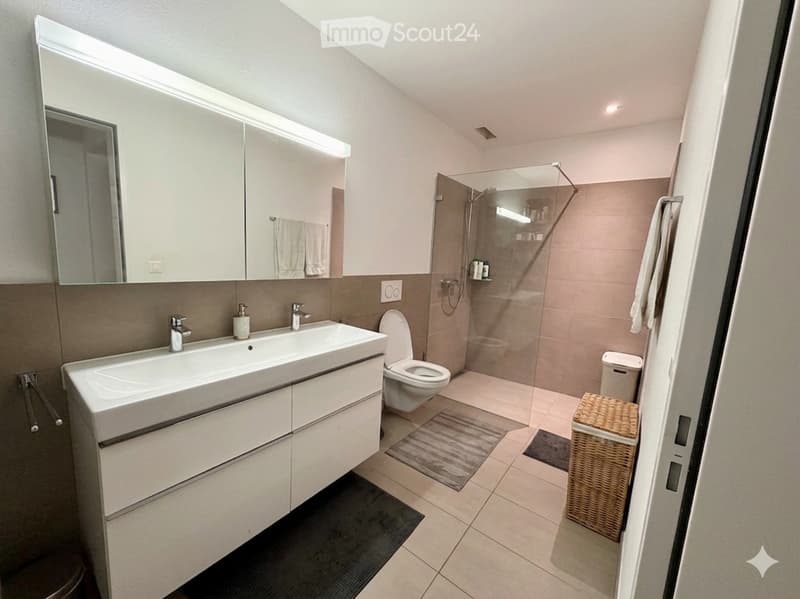 Modern bathroom with double sink, large mirror, toilet, glass-enclosed shower, towels, and wicker basket.