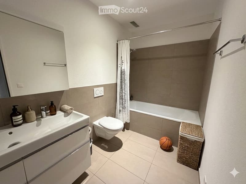 Modern bathroom with a white sink, toilet, bathtub, and shower curtain. Includes a towel, a small ball, and a wicker basket on the floor.