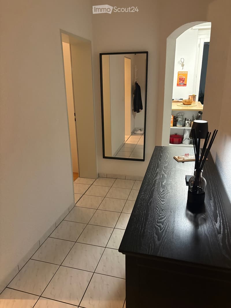 Hallway with tiled floor, a large mirror on the wall, and a black console table with decorative items on it. The hallway leads to another room with visible furniture and items.