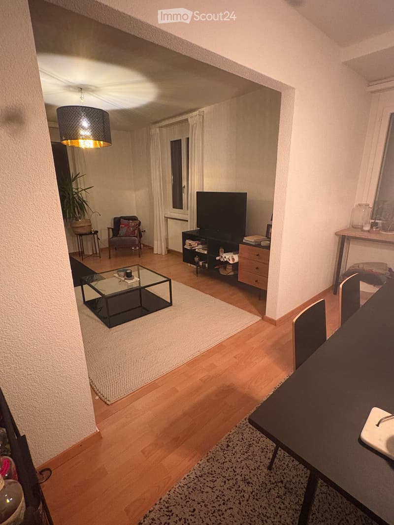 A modern living room with wooden flooring, a glass coffee table, a grey sofa, a TV stand, and a small table with chairs.