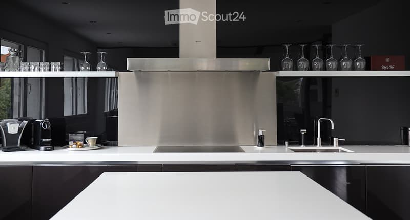 Modern kitchen with white countertops, stainless steel appliances, and black cabinets. Features a coffee machine, wine glasses, and a sink with a modern faucet.