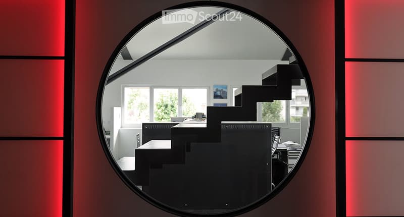 A modern, minimalist room visible through a round mirror, featuring a black chair, a table, and a screen, with large windows and artwork on the walls.