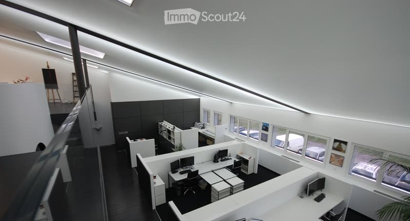 Modern office space with white desks, black chairs, multiple computer screens, and a plant near the windows.