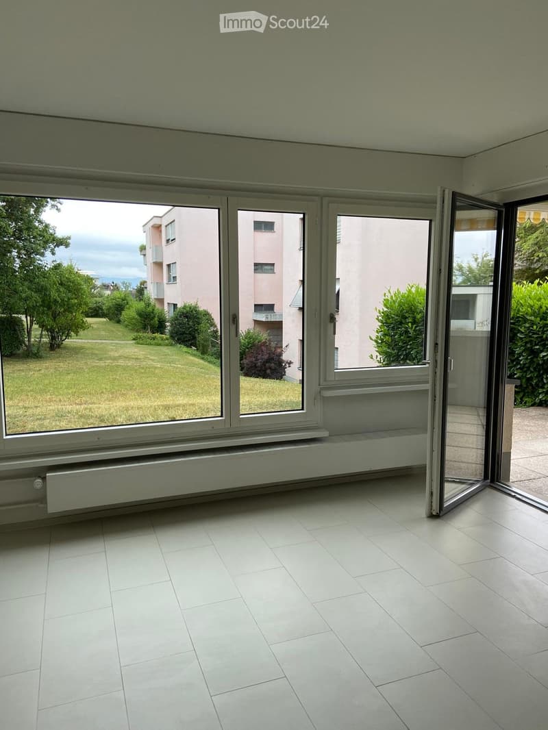 Empty room with white tiled flooring, large windows, and a radiator under the windows. The windows open to a view of a grassy area and neighboring buildings.