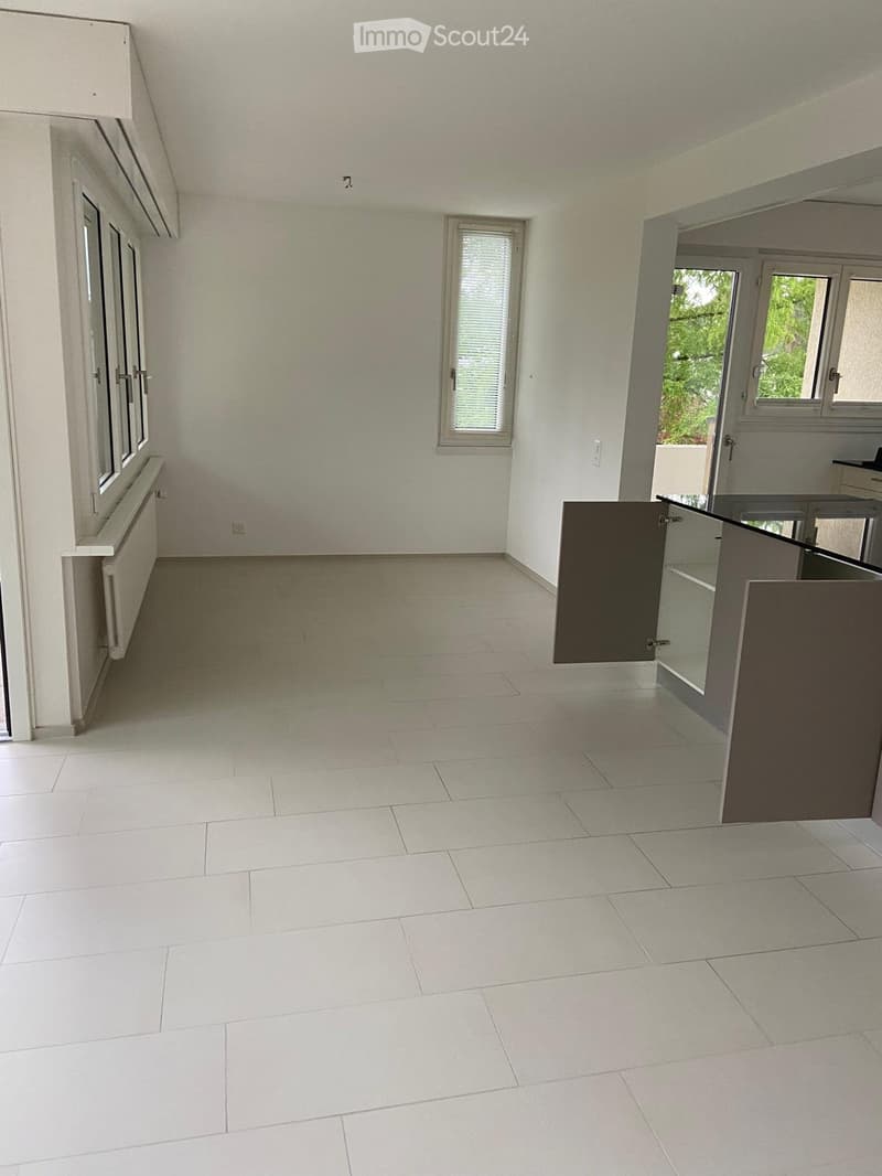 Empty room with white tiled flooring, white walls, and large windows. A small cabinet is partially visible on the right side.