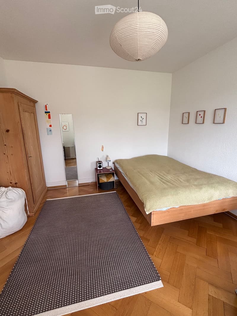 Cozy bedroom with wooden floor, bed with beige bedding, nightstand with lamp and speaker, mirror, wardrobe, and artwork on walls.