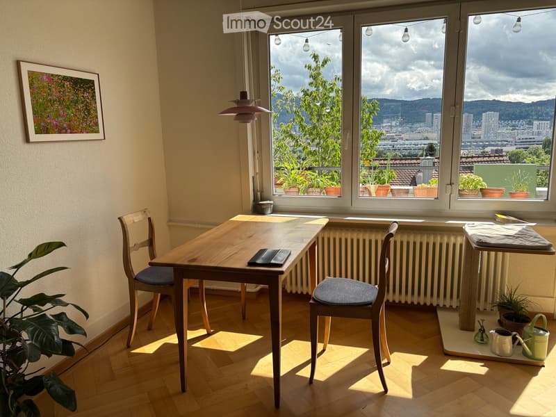 Bright room with wooden floor, large windows offering a city view, a wooden table with two chairs, a potted plant, and a picture frame on the wall.