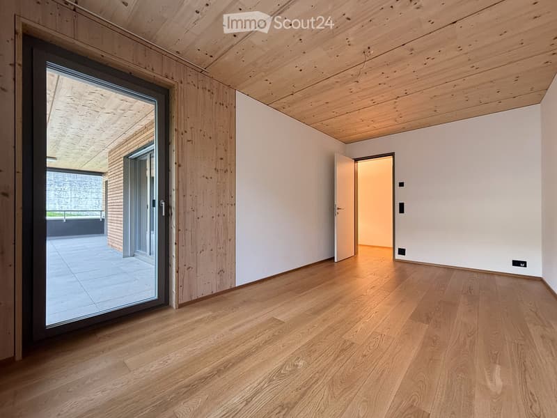 Empty hallway with light wooden flooring, white walls, and exposed wooden ceiling beams. Features a large sliding glass door leading to an outdoor area and an open doorway to another room.