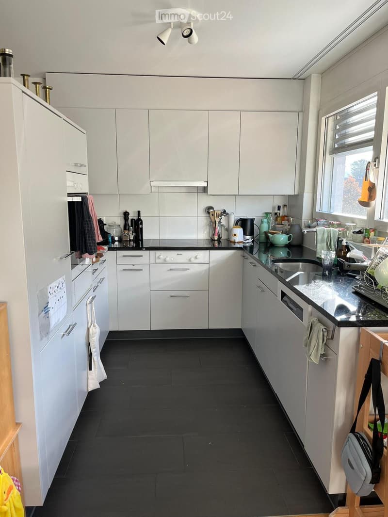Modern kitchen with white cabinets, black countertops, and black tiled flooring. Visible appliances include an oven, dishwasher, and sink. Various kitchen items such as bottles, cups, and a kettle are on the countertops.