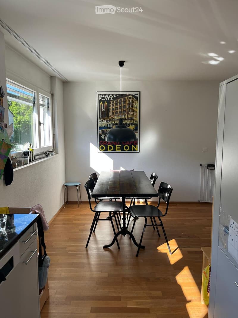 Bright, open room with wooden flooring, a central dining table with chairs, a pendant light, and a poster on the wall. A small stool is near the window, and a refrigerator is visible on the right side.