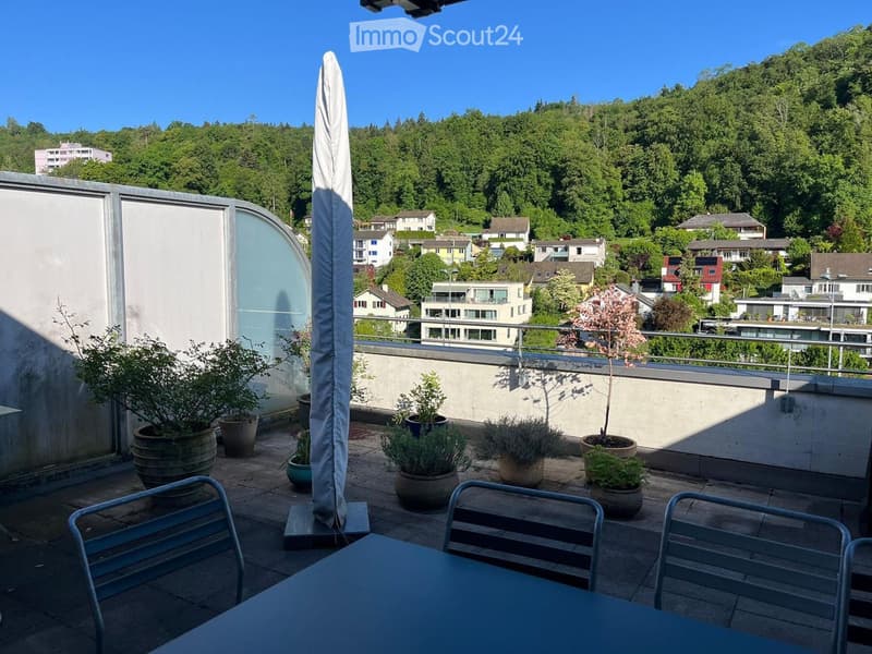 Balcony with a table, chairs, umbrella, and several potted plants, overlooking a green, hilly landscape with houses.