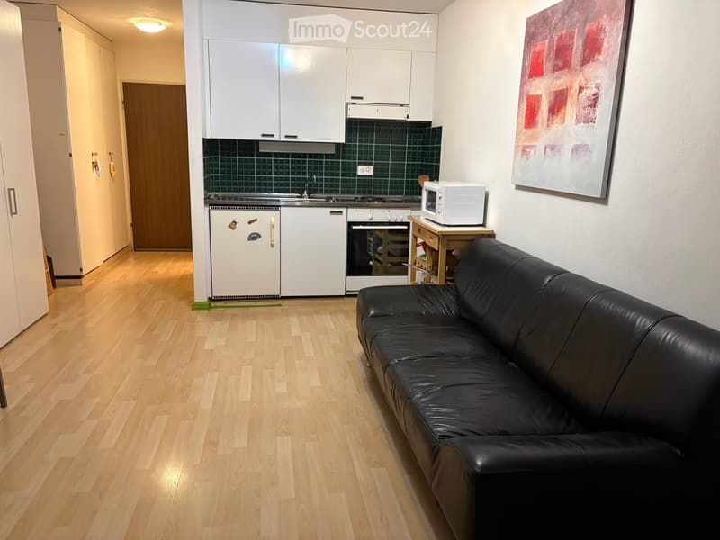 A modern, furnished room with a black leather sofa, a small kitchen area featuring white cabinets, a microwave, and a small table, along with a painting on the wall.