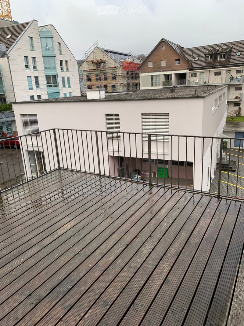 A balcony with wooden floor and metal railings, overlooking a street with parked cars and buildings.