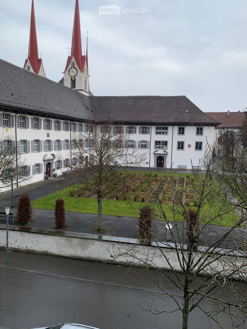 Large white building, brown roof, two red steeples, clock, windows, grassy garden, paved path, two benches, street light, cloudy sky