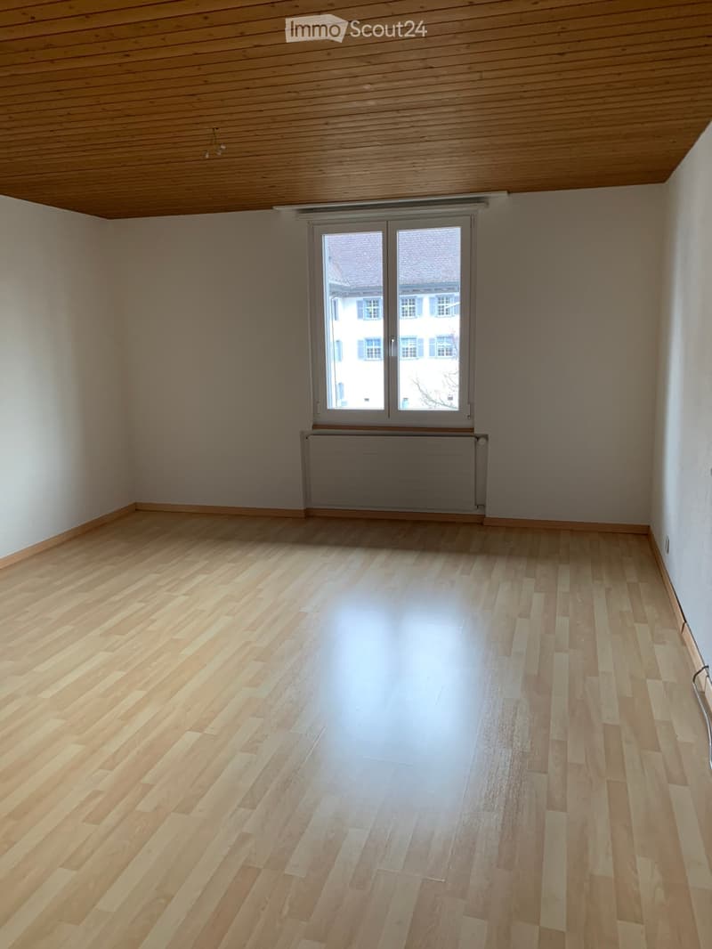 Spacious room with wooden floor, white walls, large window, wooden ceiling, and a radiator.