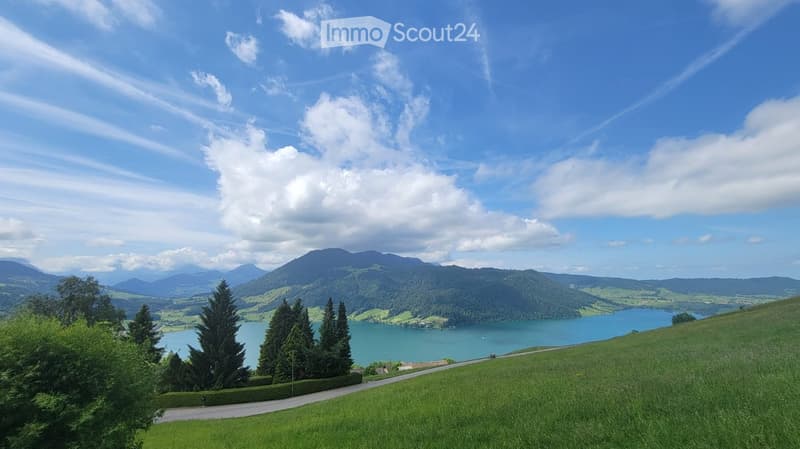 A scenic outdoor view featuring a lake surrounded by lush green hills and mountains under a partly cloudy sky. The foreground shows a grassy slope with a winding road leading towards the lake.